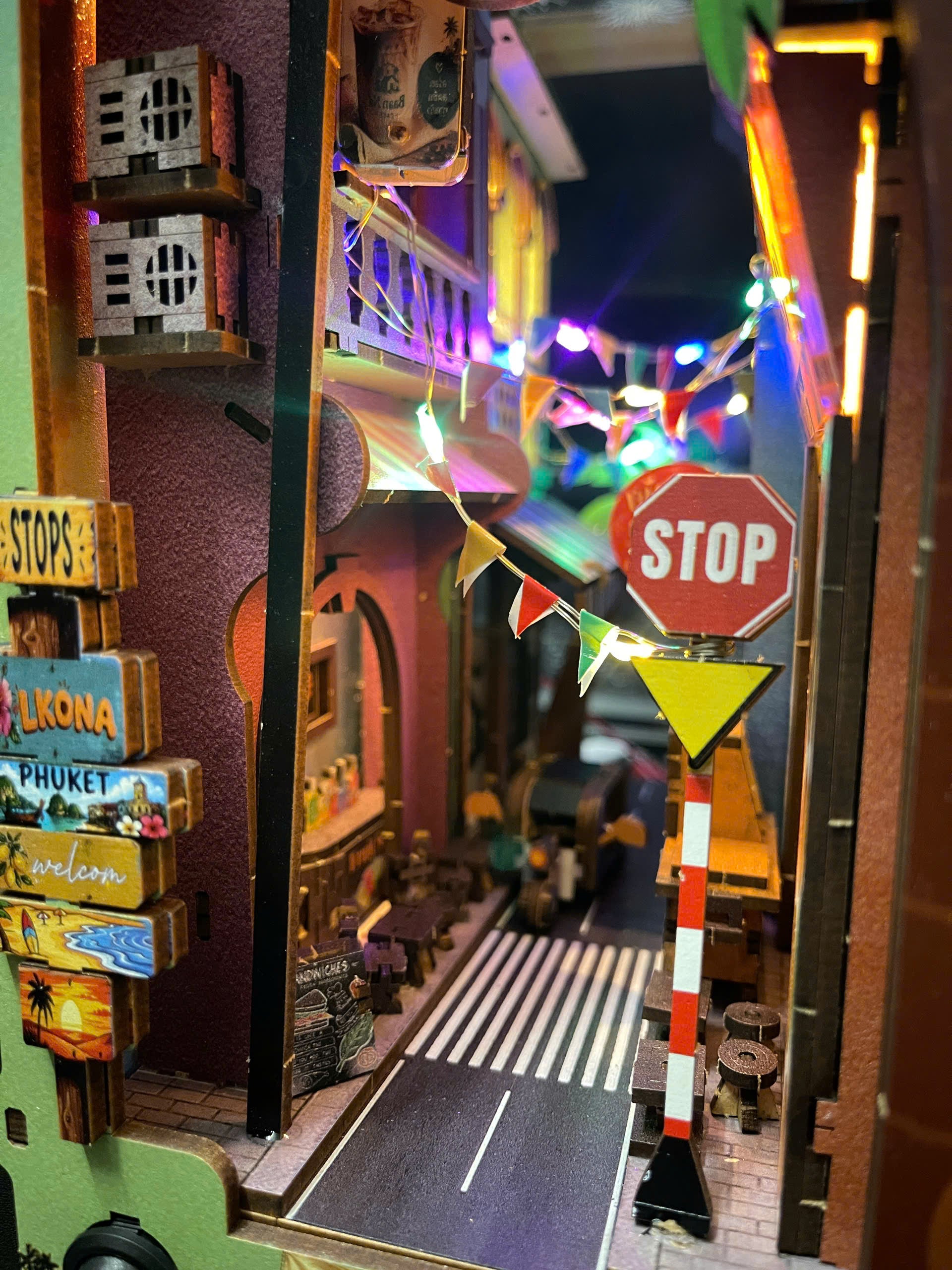 A miniature diorama of a street scene with toy cars, colorful string lights, a stop sign, and decorative signs including 'Lomapa Phuket' and 'Welcom'.