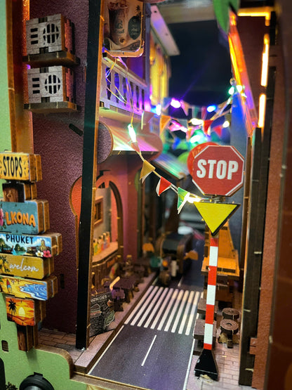 A miniature diorama of a street scene with toy cars, colorful string lights, a stop sign, and decorative signs including 'Lomapa Phuket' and 'Welcom'.