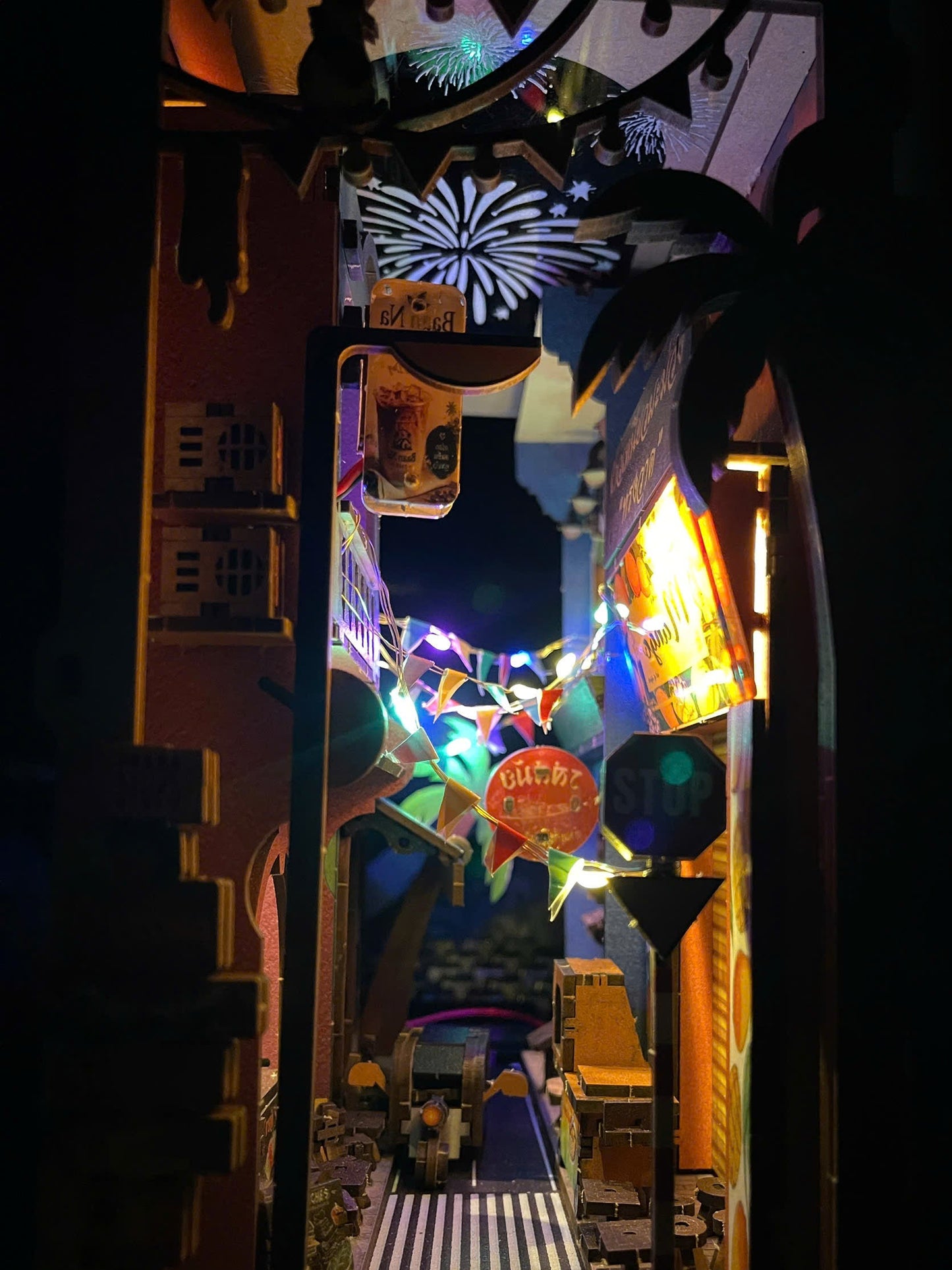 A miniature diorama of a nighttime street scene with glowing multicolored signs, palm trees, fireworks, a crosswalk, and tiny vehicles inside a dark, illuminated display.