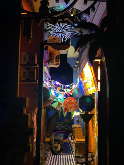 A miniature diorama of a nighttime street scene with glowing multicolored signs, palm trees, fireworks, a crosswalk, and tiny vehicles inside a dark, illuminated display.
