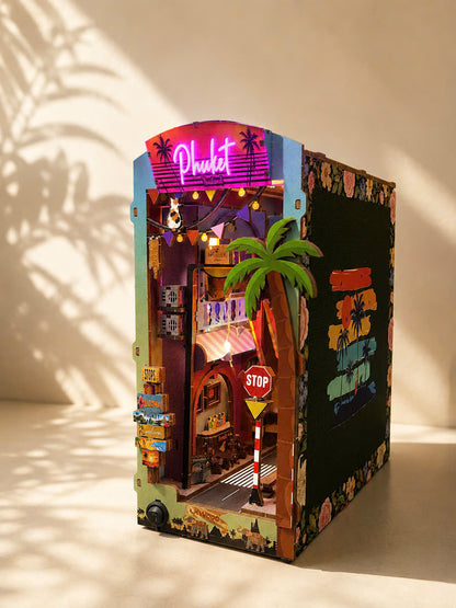 A miniature cardboard city building kit styled as a vibrant street scene with a 'Pleasant' neon sign, palm tree, and colorful retro storefront details, photographed on a warm beige background with leaf shadows.